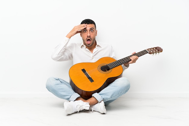 Jonge man zittend op de vloer met gitaar met verrassing en geschokt gelaatsuitdrukking