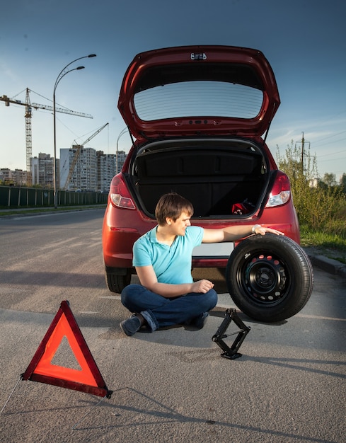 Jonge man zittend op de weg en leunend tegen kapotte auto