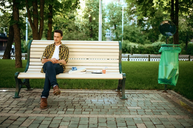 Jonge man zittend op een bankje tussen het afval in het park, vrijwilligerswerk. ecologische restauratie, ecologische levensstijl, ecologische zorg, milieureinigingsconcept