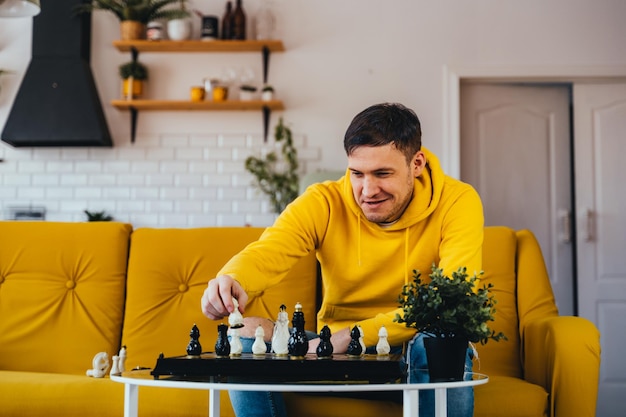 Jonge man zittend op een gele bank en schaken in de kamer Man speelt in logisch bordspel met zichzelf