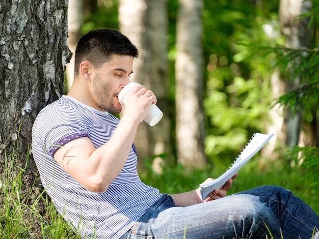 Jonge man zittend op een gras in het park een boek lezen en koffie drinken