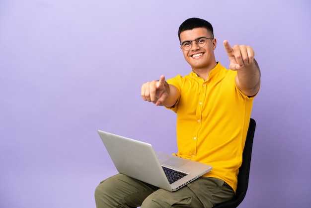 Jonge man zittend op een stoel met laptop wijst vinger naar je terwijl hij lacht