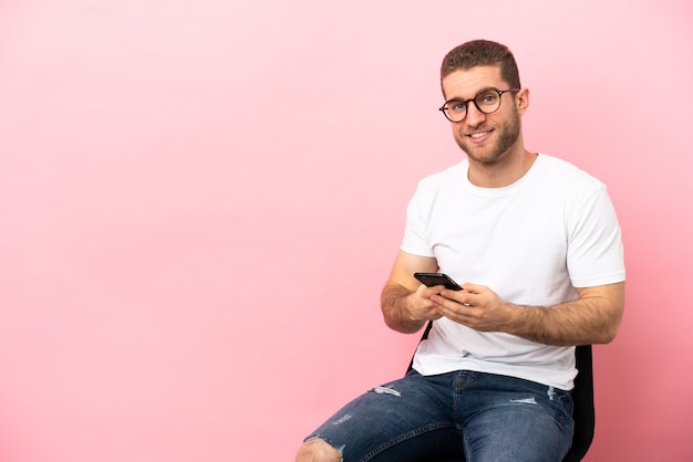 Jonge man zittend op een stoel over geïsoleerde roze achtergrond die een bericht verzendt met de mobiel