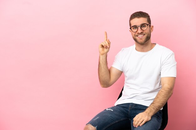 Jonge man zittend op een stoel over geïsoleerde roze achtergrond die een vinger toont en optilt in teken van het beste