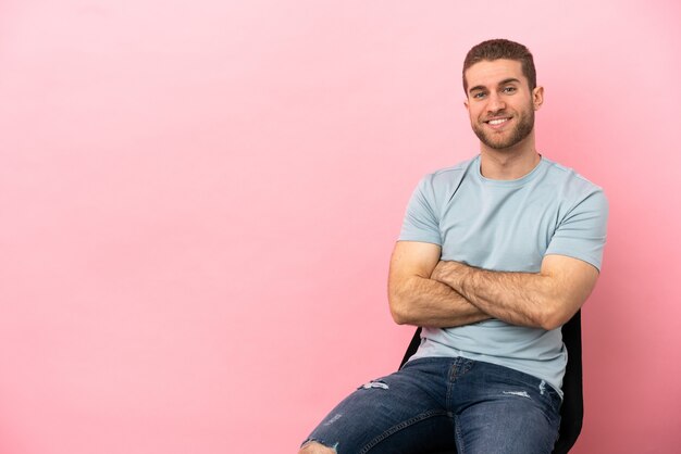 Jonge man zittend op een stoel over geïsoleerde roze achtergrond met armen gekruist en vooruit kijkend