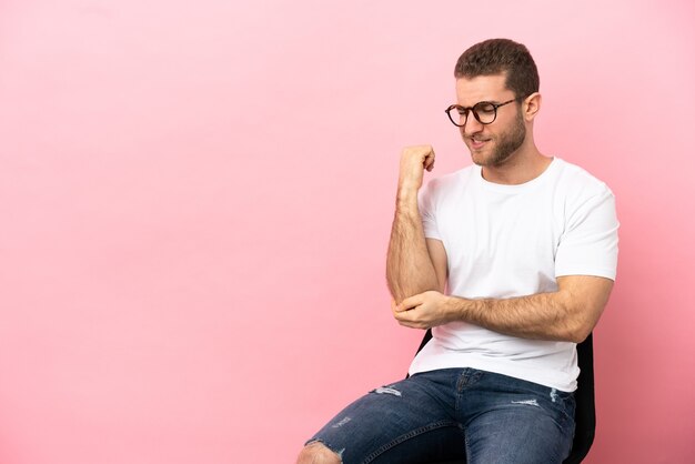 Jonge man zittend op een stoel over geïsoleerde roze achtergrond met pijn in de elleboog