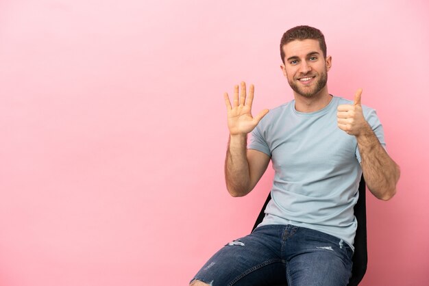 Jonge man zittend op een stoel over geïsoleerde roze achtergrond zes tellen met vingers