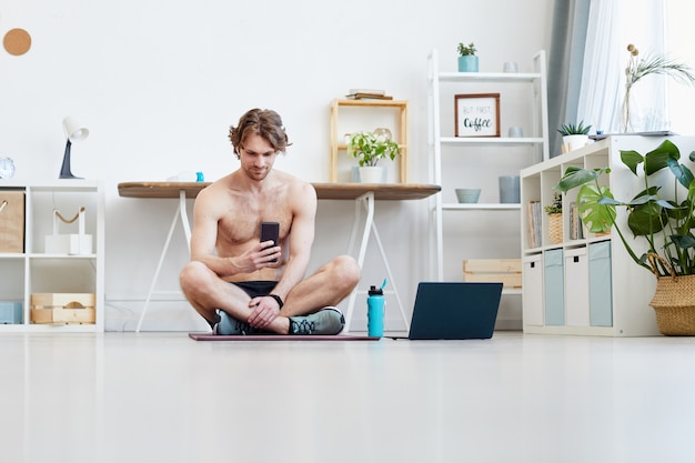 Jonge man zittend op oefeningsmat en online praten op zijn mobiele telefoon tijdens het rusten na sporttraining thuis