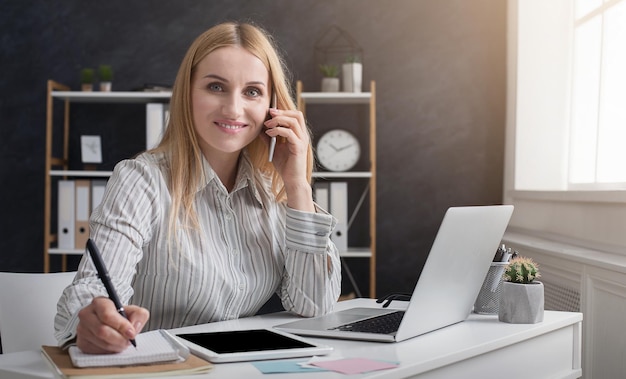 Jonge manager belt op mobiele telefoon tijdens het schrijven van notities op kladblok op kantoor