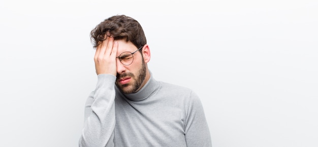 Jonge manager man op zoek slaperig, verveeld en geeuwen, met een hoofdpijn en een hand die de helft van het gezicht over witte muur