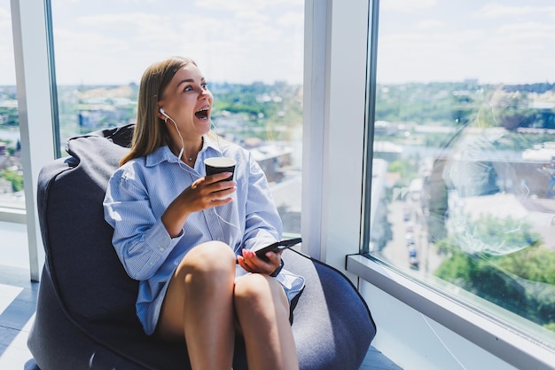 Jonge managervrouw met koptelefoon die aan de telefoon praat en koffie drinkt terwijl ze in een moderne coworking-ruimte zit Freelance meisje dat op afstand online werkt