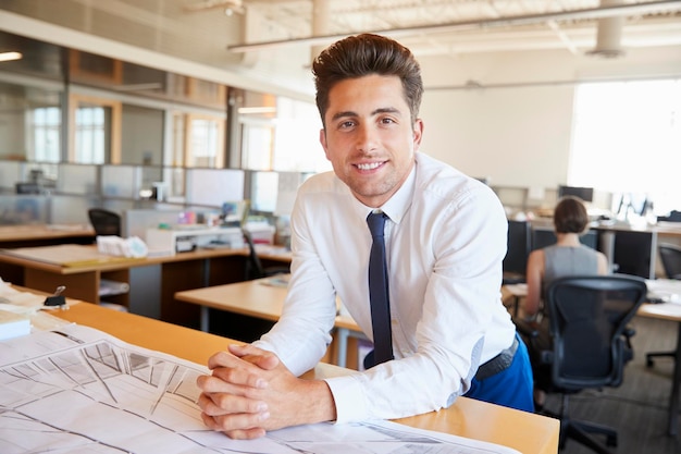 Jonge mannelijke architect leunend op bureau glimlachend naar camera