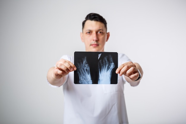 Jonge mannelijke arts in een wit chirurgisch pak houdt een röntgenfoto van de beenbotten van de patiënt vast en onderzoekt deze. Geïsoleerd op een witte achtergrond