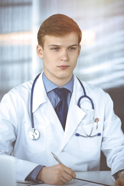 Jonge mannelijke arts zitten en werken met klembord van medicatie geschiedenis record in zonnige kliniek op zijn werkplek. Geneeskunde concept.