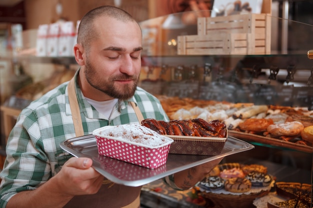 Jonge mannelijke bakker die heerlijke versgebakken cakes ruikt die hij bij zijn bakkerijwinkel verkoopt