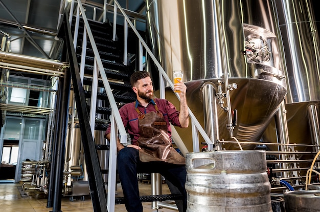 Jonge mannelijke brouwer in leren schort bij moderne brouwerijfabriek