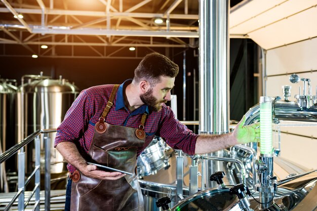 Jonge mannelijke brouwer in leren schort die toezicht houdt op het proces van bierfermentatie bij moderne brouwerijfa...