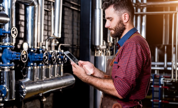 Jonge mannelijke brouwer in leren schort die toezicht houdt op het proces van bierfermentatie bij moderne brouwerijfa...