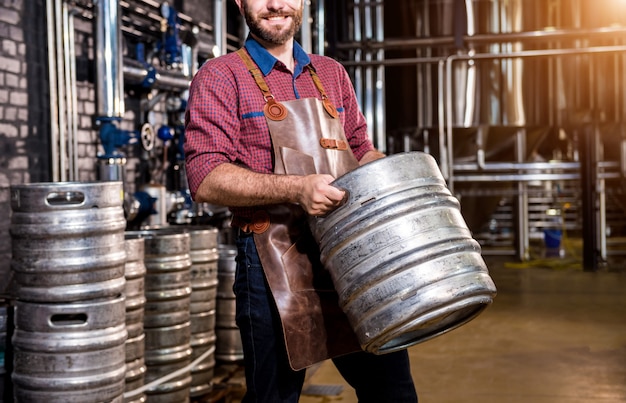 Jonge mannelijke brouwer in leren schort houdt vat met ambachtelijk bier vast in moderne brouwerijfabriek