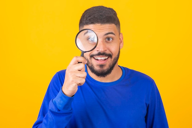 Jonge mannelijke detective die vergrootglas met het rechteroog vasthoudt terwijl hij geïsoleerd voor de camera staat