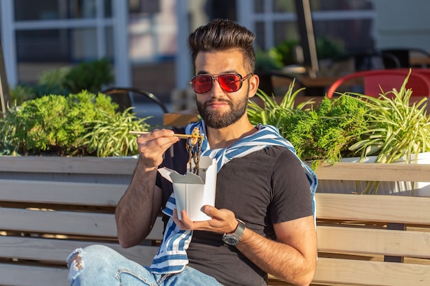 Jonge mannelijke hipster met een snor en baard eet chinese noedels met houten eetstokjes uit een witte lunchbox. Aziatische keuken concept.