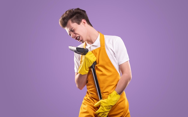 Jonge mannelijke huishoudster in gele schort en handschoenen zingen in stofzuiger met gesloten ogen tijdens huishoudelijke routine tegen violette achtergrond