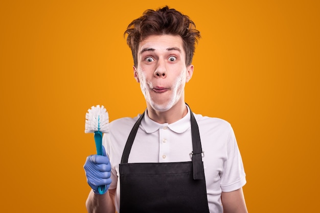 Jonge mannelijke huishoudster met toiletborstel grimassen en camera kijken tegen gele achtergrond