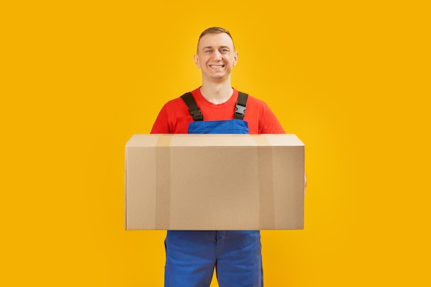 Jonge mannelijke koerier in blauw uniform houdt groot pakket in zijn handen Bezorger met papieren doos op gele achtergrond Kopieer ruimte mock up