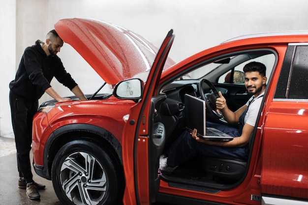 Jonge mannelijke monteur met behulp van laptop zittend in rode auto en zijn collega met tablet onder motorkap