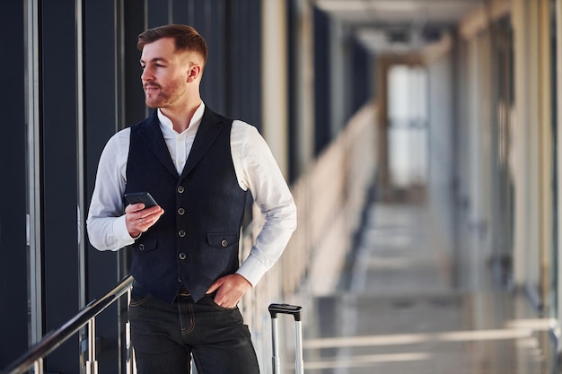 Jonge mannelijke passagier in elegante formele kleding staat in de luchthavenhal met bagage en telefoon in de hand.