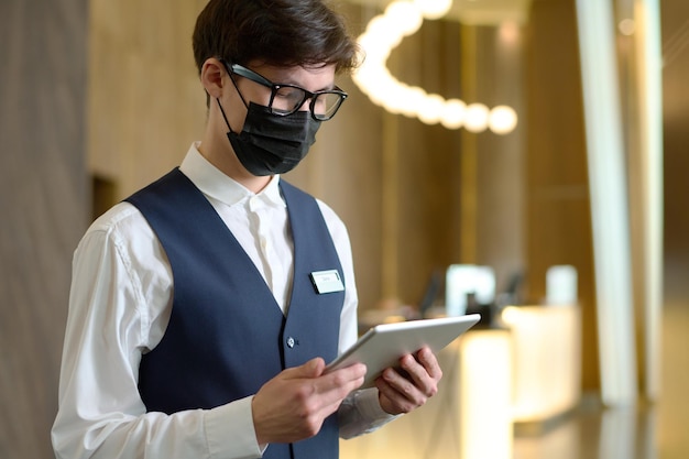 Jonge mannelijke receptioniste van modern hotel in uniform en masker met tablet