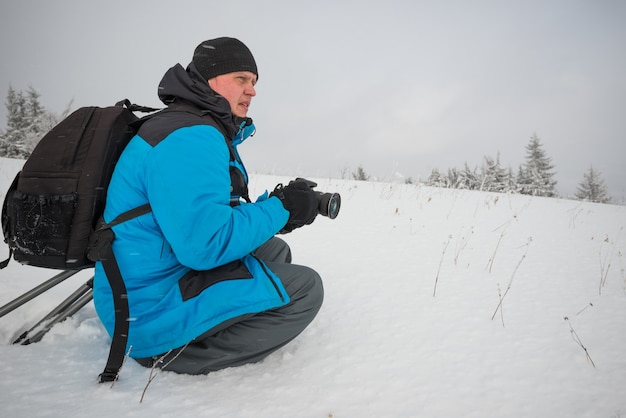 Jonge mannelijke reiziger met rugzak maakt foto's van een prachtige hoge besneeuwde dennenboom in een hoge sneeuwjacht tegen de achtergrond van mist op een ijzige winterdag