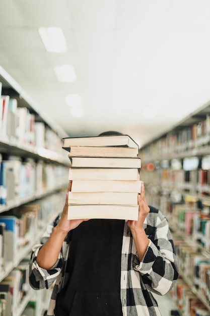 Jonge mannelijke student die zijn gezicht bedekt door de stapel boeken in het midden van de gangpadbibliotheek