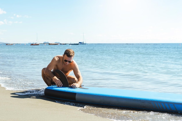 Jonge mannelijke surfer die stand-up paddleboard op een strand opzet