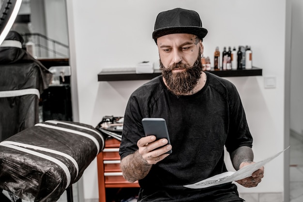 Jonge mannelijke tatoeëerder met baard met telefoon en schets zittend op de Bank in workshop plaats.