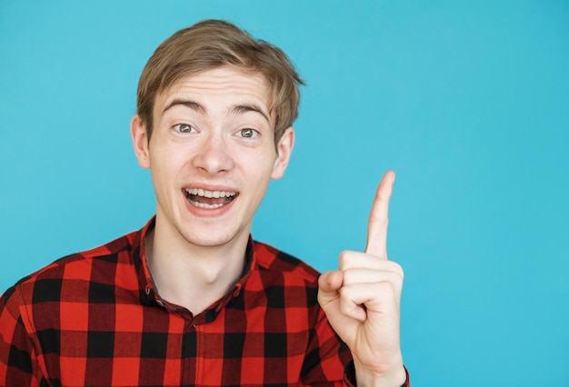 Jonge mannelijke tiener in rood shirt op blauwe achtergrond wijst vinger naar kopieerruimte heeft een idee