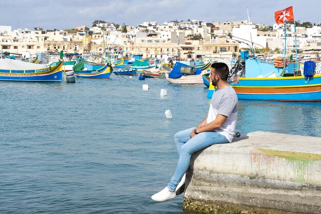 Jonge mannelijke toerist zit en op de achtergrond de zee vol kleurrijke boten bij Marsaxlokk in Malta