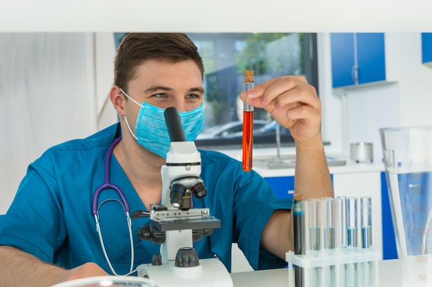 Jonge mannelijke wetenschapper in uniform die een masker draagt, houdt een reageerbuis met rode vloeistof vast en doet wat onderzoek in een laboratorium. Gezondheidszorg en biotechnologie concept