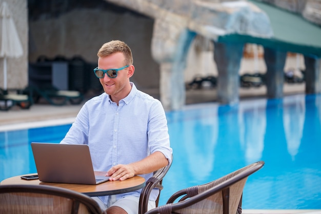 Jonge mannelijke zakenman in zonnebril werkt op een laptop zittend aan een tafel bij het zwembad. Afstandswerk. Freelancer