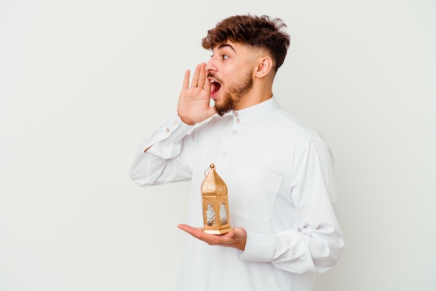 Jonge marokkaanse man die een typische arabische kleding draagt die een ramadan-lamp houdt die op wit wordt geïsoleerd schreeuwen en palm dichtbij geopende mond houden.