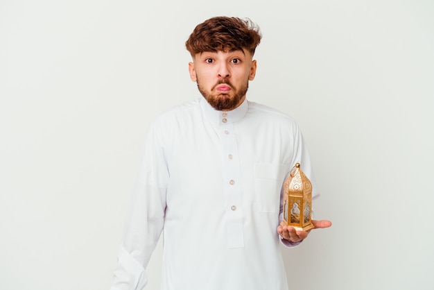 Jonge Marokkaanse man die een typische Arabische kleding draagt die een ramadan-lamp vasthoudt die op wit wordt geïsoleerd, haalt zijn schouders op en opent verwarde ogen.