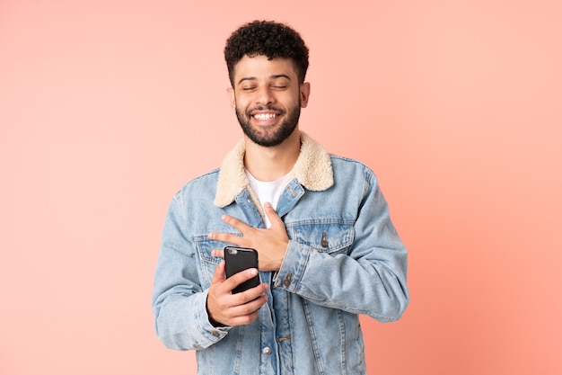 Jonge Marokkaanse man met behulp van mobiele telefoon geïsoleerd op roze muur veel glimlachen