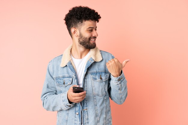 Jonge Marokkaanse man met behulp van mobiele telefoon geïsoleerd op roze muur wijzend naar de zijkant om een product te presenteren