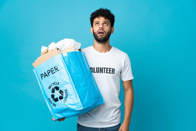 Jonge Marokkaanse man met een recyclingzak vol papier om te recyclen over geïsoleerde op zoek omhoog en met verbaasde uitdrukking