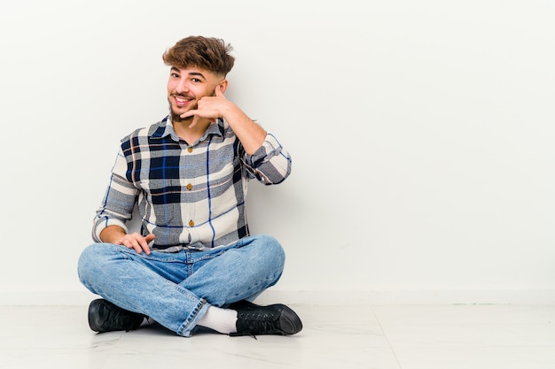 Jonge Marokkaanse man zittend op de vloer geïsoleerd op een witte muur met een mobiele telefoongesprek gebaar met vingers.
