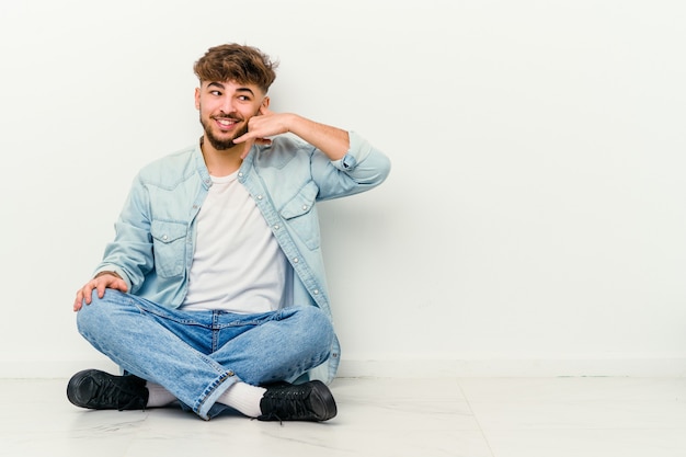 Jonge Marokkaanse man zittend op de vloer geïsoleerd op wit met een mobiele telefoongesprek gebaar met vingers.