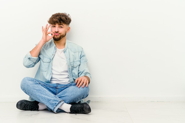 Jonge Marokkaanse man zittend op de vloer geïsoleerd op wit met vingers op lippen een geheim bewaren.