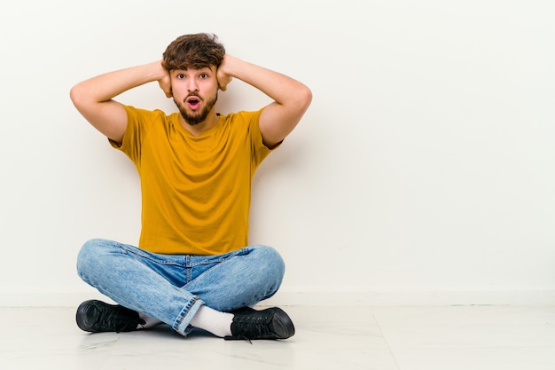 Jonge Marokkaanse man zittend op de vloer geïsoleerd op wit oren bedekken met handen proberen niet te hard geluid te horen.