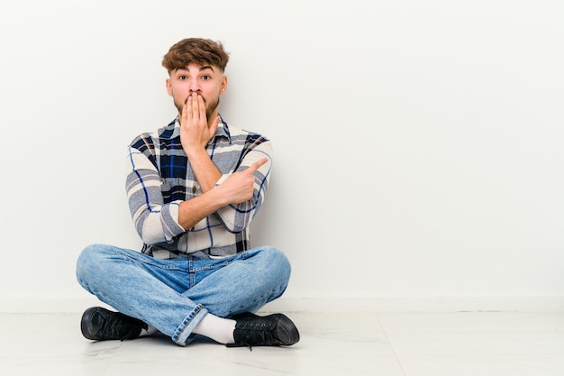 Jonge Marokkaanse man zittend op de vloer geïsoleerd op wit wijzend naar de zijkant