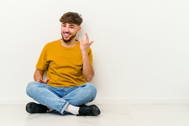 Jonge Marokkaanse man zittend op de vloer met rock gebaar met vingers